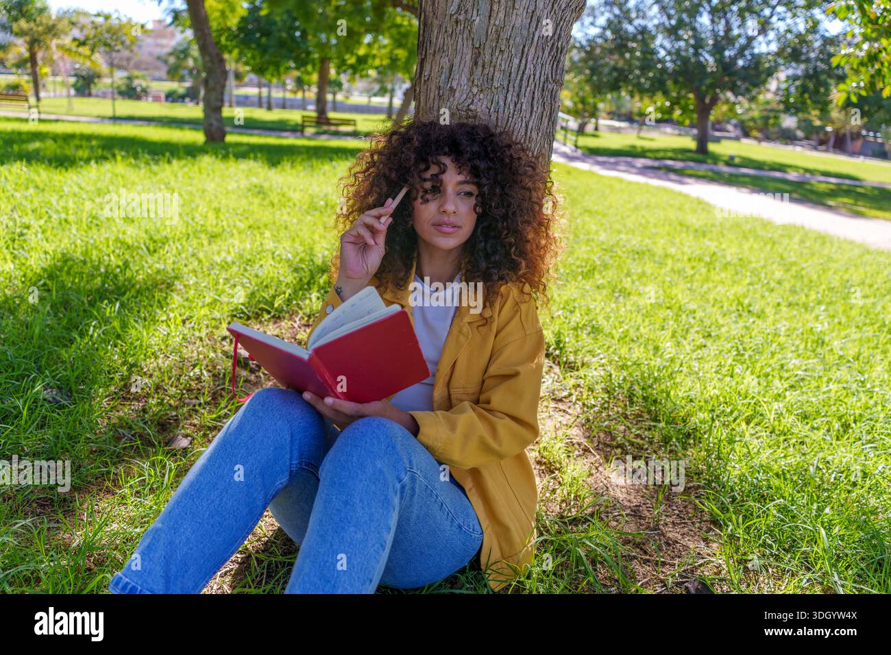 Woman with curly hair sitting on grass leaning on a tree trunk, holding a red notebook and pencil, thinking and creating Stock Photo
