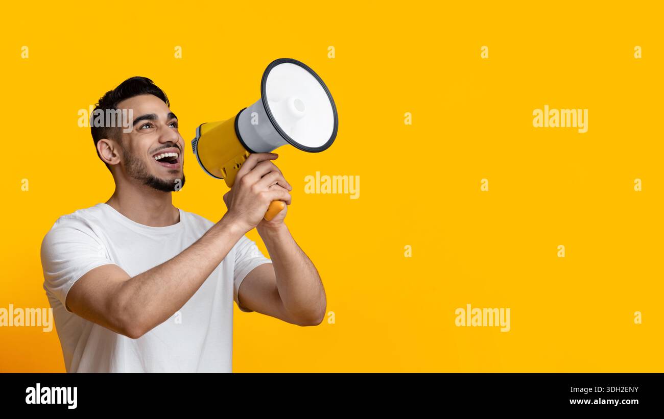 Excited arab guy speaking loud, using megaphone on yellow Stock Photo