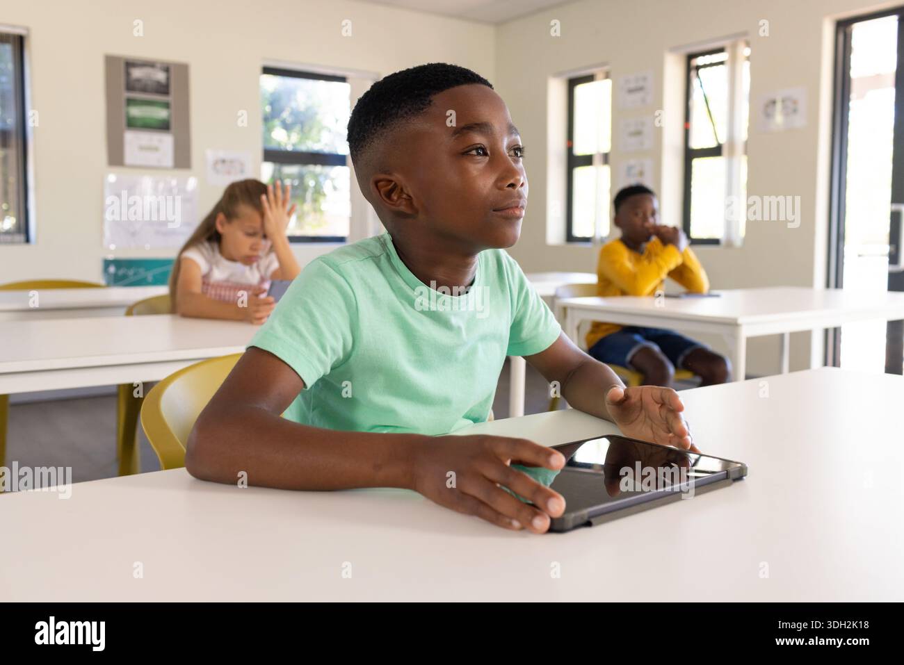 Diverse youth classmates learning at white desks under windows, boy in light green tee using tablet Stock Photo