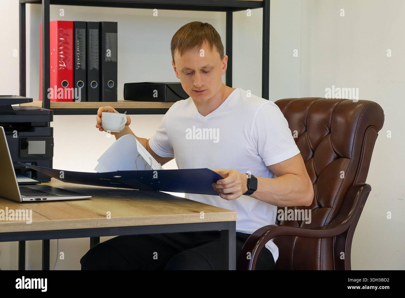 Seated man holds white cup, gazing puzzled at open blue folder with documents on desk, surrounded by office equipment. Stock Photo