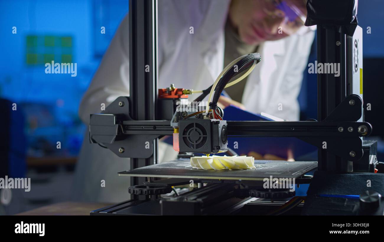 Engineer in lab coat and safety glasses adjusts 3D printer while creating a prototype in a modern lab focused on advanced technology. Stock Photo