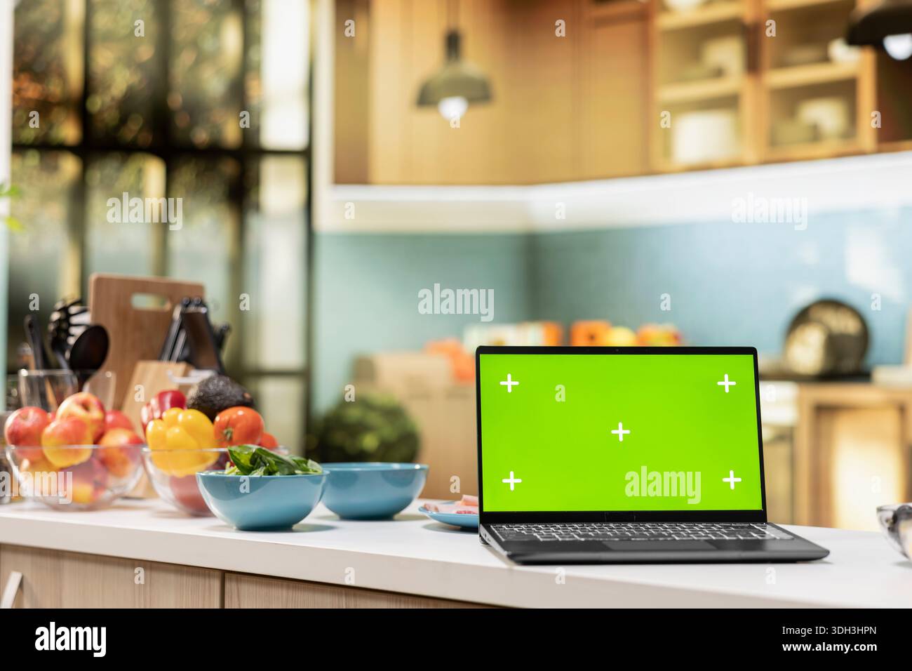 Green screen on a laptop placed on empty table in modern kitchen space decorated with modern appliances, cooking essentials and kitchenware. Comfortable dining room with light. Stock Photo