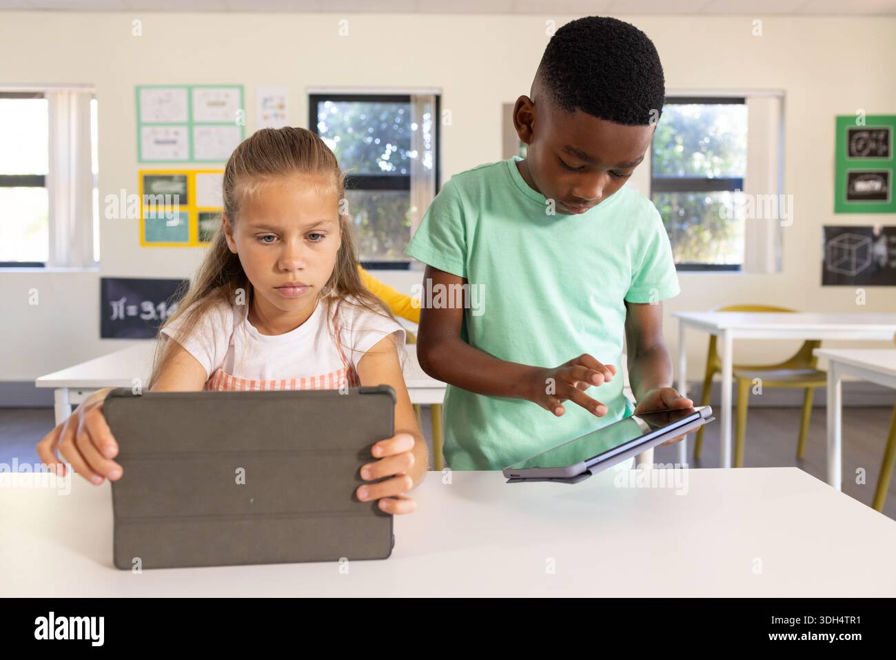 Diverse students sitting at white table in bright classroom using tablets in folio cases Stock Photo