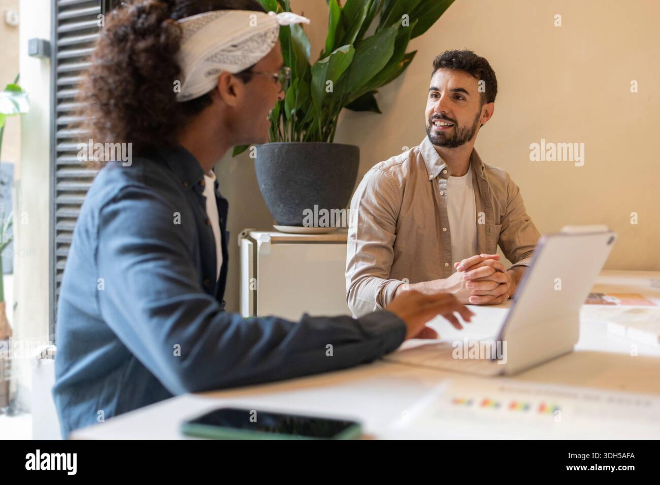 Two professionals engaging in a collaborative discussion at a coworking space. One is using a tablet while both are surrounded by a modern, creative e Stock Photo