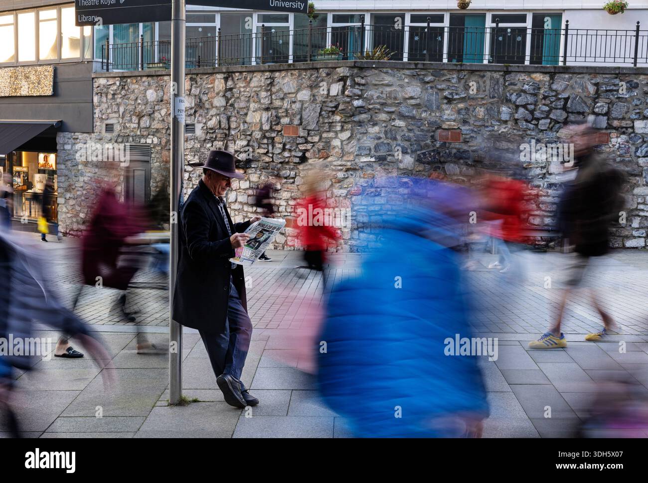 A man in a black coat and hat reading a newspaper in the hustle and bustleof a busy street. Stock Photo