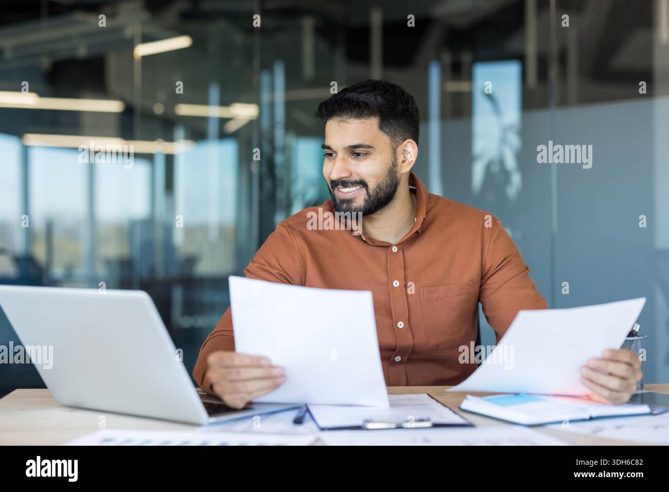 Young indian businessman at a modern office desk, smiling while reviewing reports and laptop, planning finances and strategy with confident, focused professionalism and productivity Stock Photo