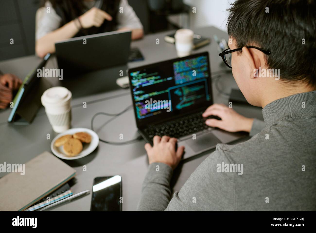 Young Asian Man Writing Code on Laptop While Collaborating with Colleagues Stock Photo