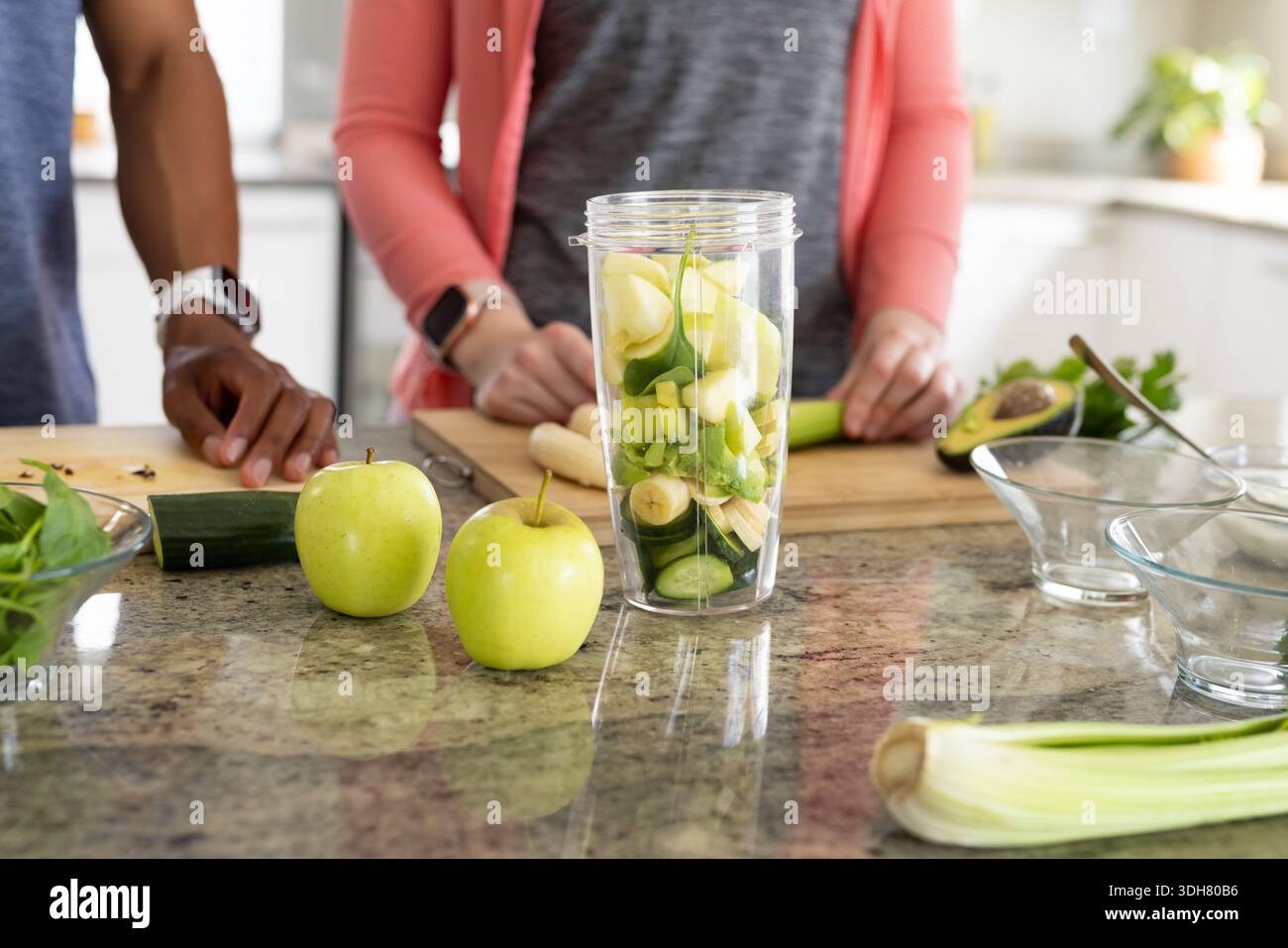 Diverse couple preparing green smoothie at kitchen counter with blender wearing smartwatches Stock Photo