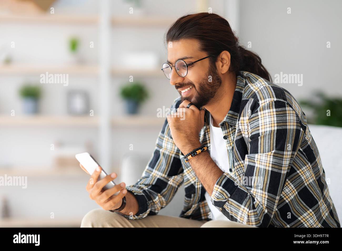 Cheerful long-haired middle-eastern guy hipster sitting on couch, using smartphone Stock Photo