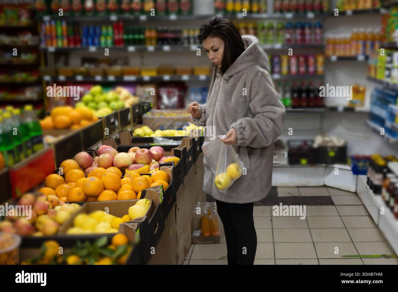 A young woman wearing a gray coat stands at a produce section in a grocery store. She is carefully selecting yellow apples and placing them into a clear plastic bag. Stock Photo