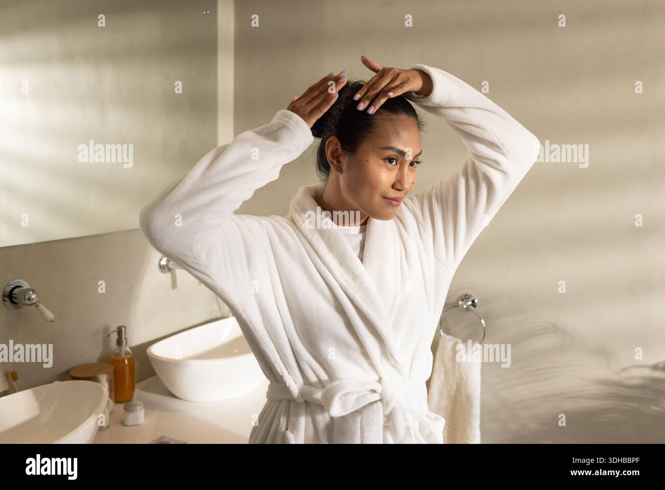 Asian woman standing at vanity in bathroom in white robe, adjusting hair into bun by mirror Stock Photo