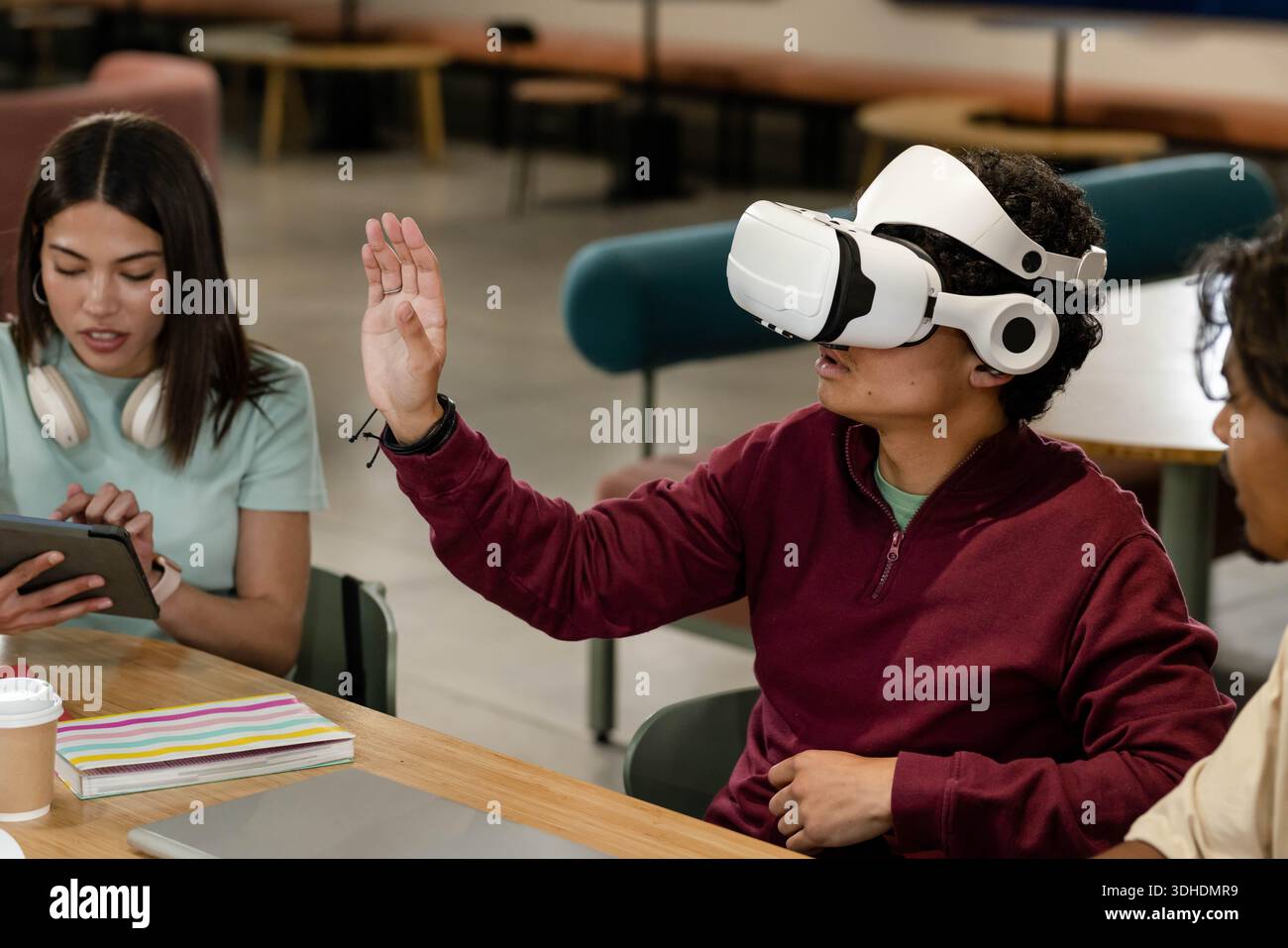 African American students sitting in campus lounge around table using VR headset, tablet, laptop Stock Photo