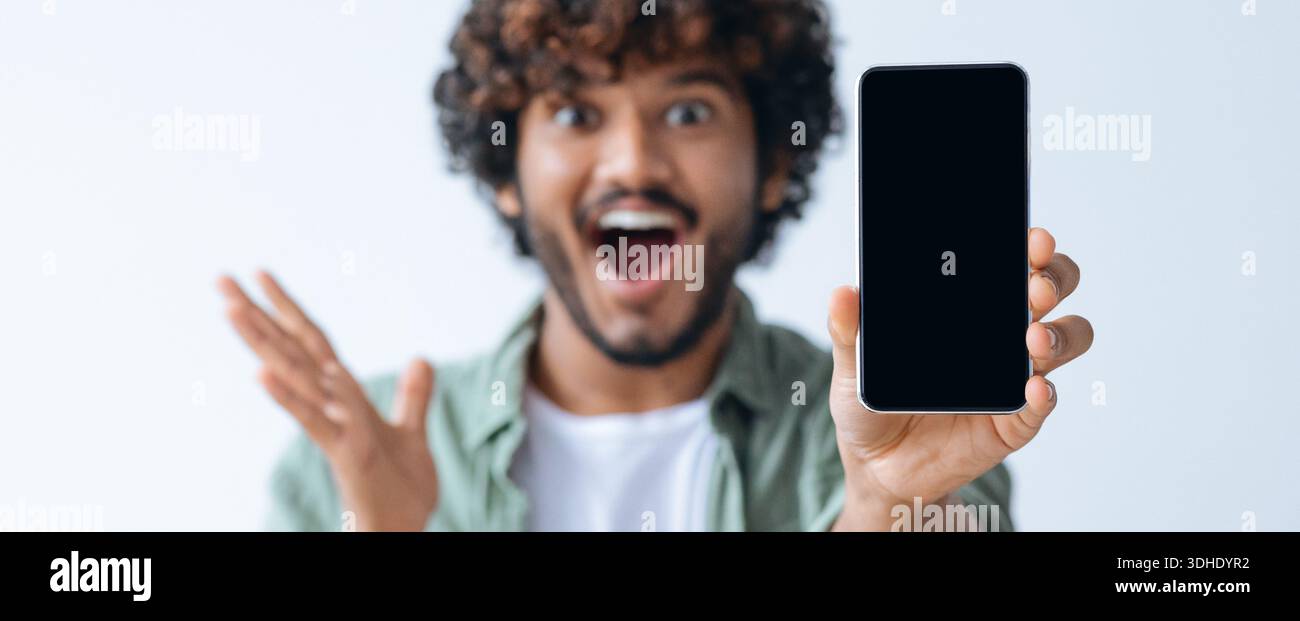 Excited Young Man Shows Smartphone in Bright Setting While Smiling and Gesturing Stock Photo