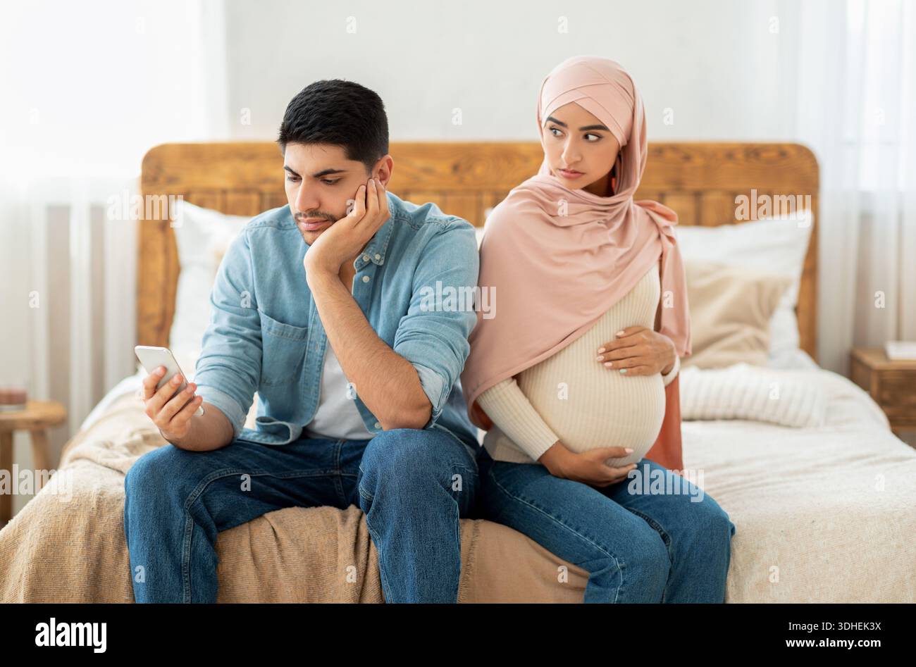 Young pregnant muslim woman offended to her husband busy with smartphone, arab lady in hijab feeling lonely Stock Photo