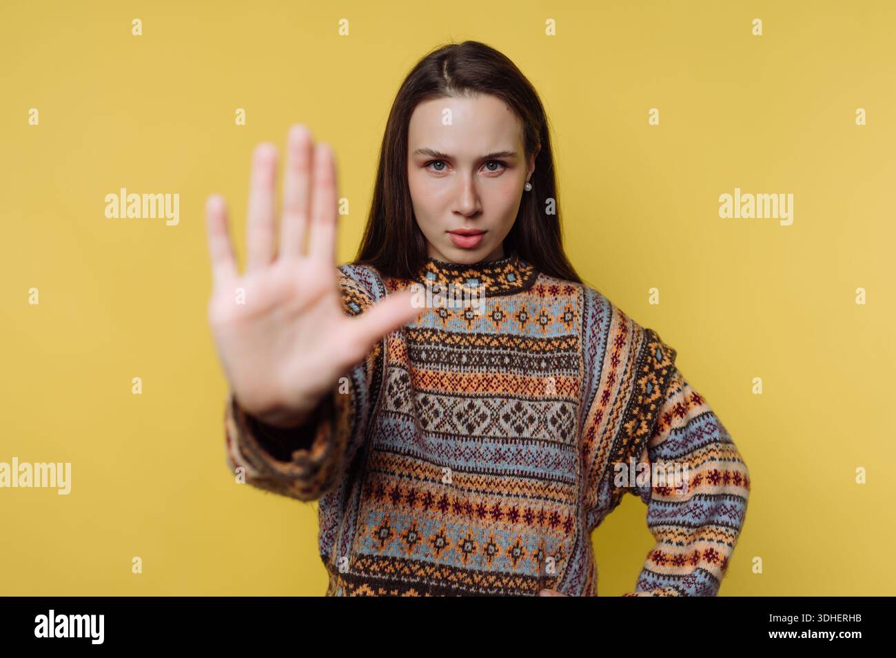 Woman in patterned sweater making stop gesture on yellow background Stock Photo