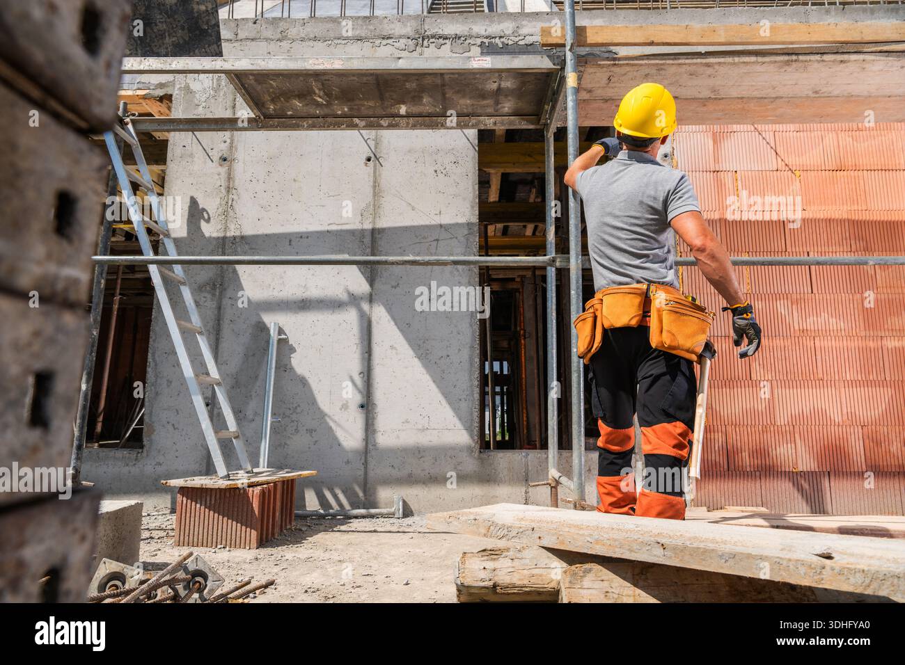 A construction worker stands on a site working on wall materials while wearing a yellow hard hat and safety gear. Stock Photo