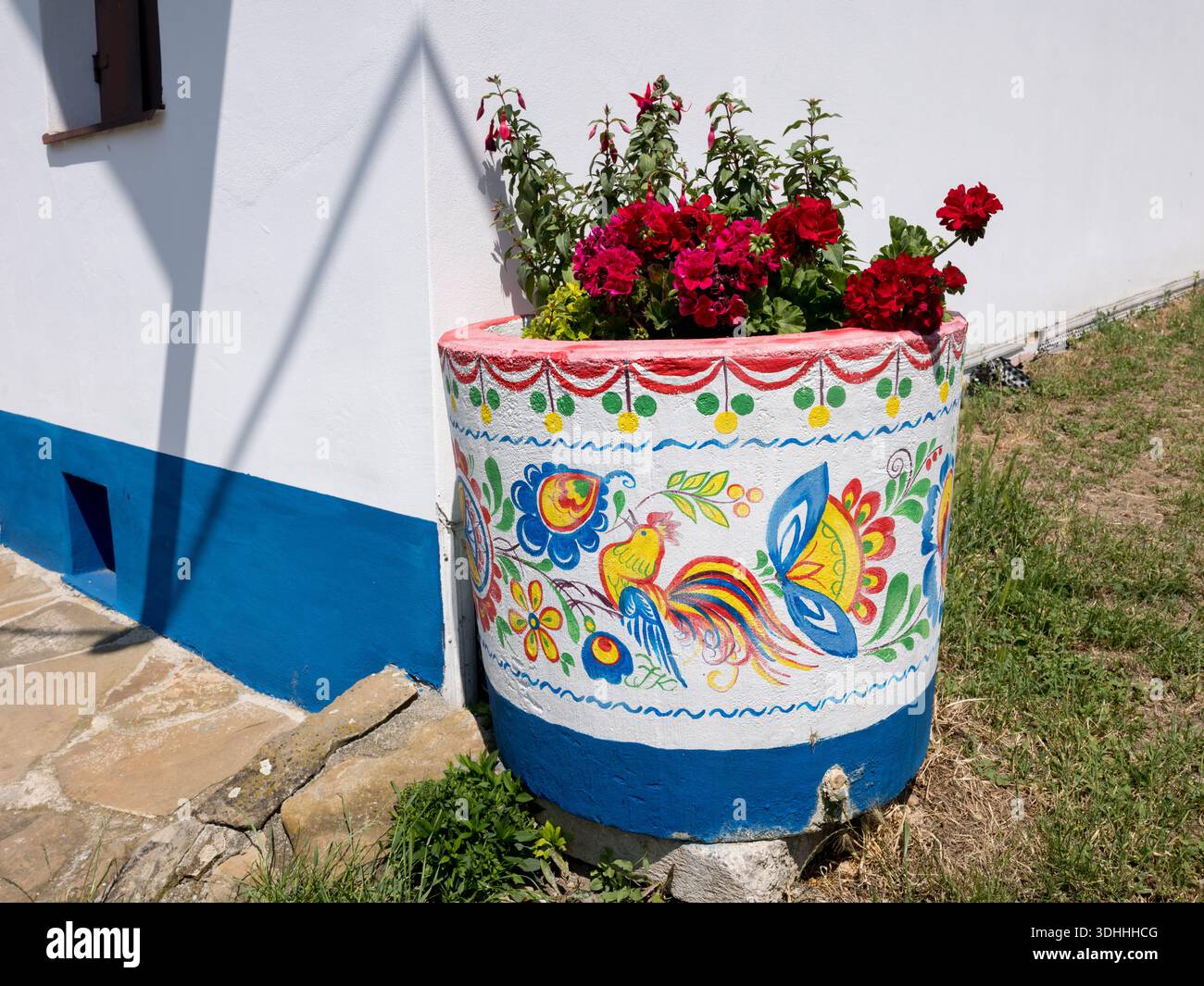 A large concrete flowerpot with traditional Czech folk painting, featuring a rooster and floral motifs, standing by a white and blue house wall Stock Photo
