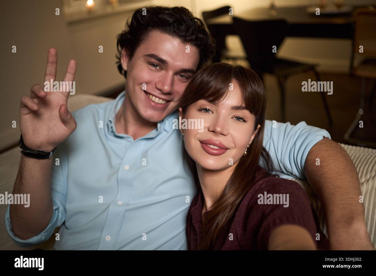 Portrait of Caucasian young adult man smiling and making peace sign, while sitting next to Caucasian young adult woman, taking selfie together on sofa in home interior Stock Photo
