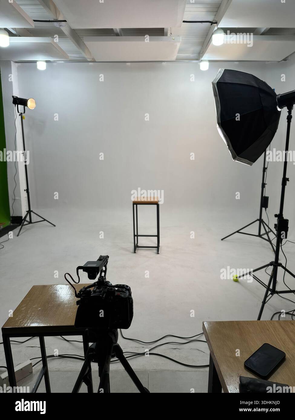 Empty photography studio setup with lights, softbox, camera on tripod and stool on white backdrop. Stock Photo