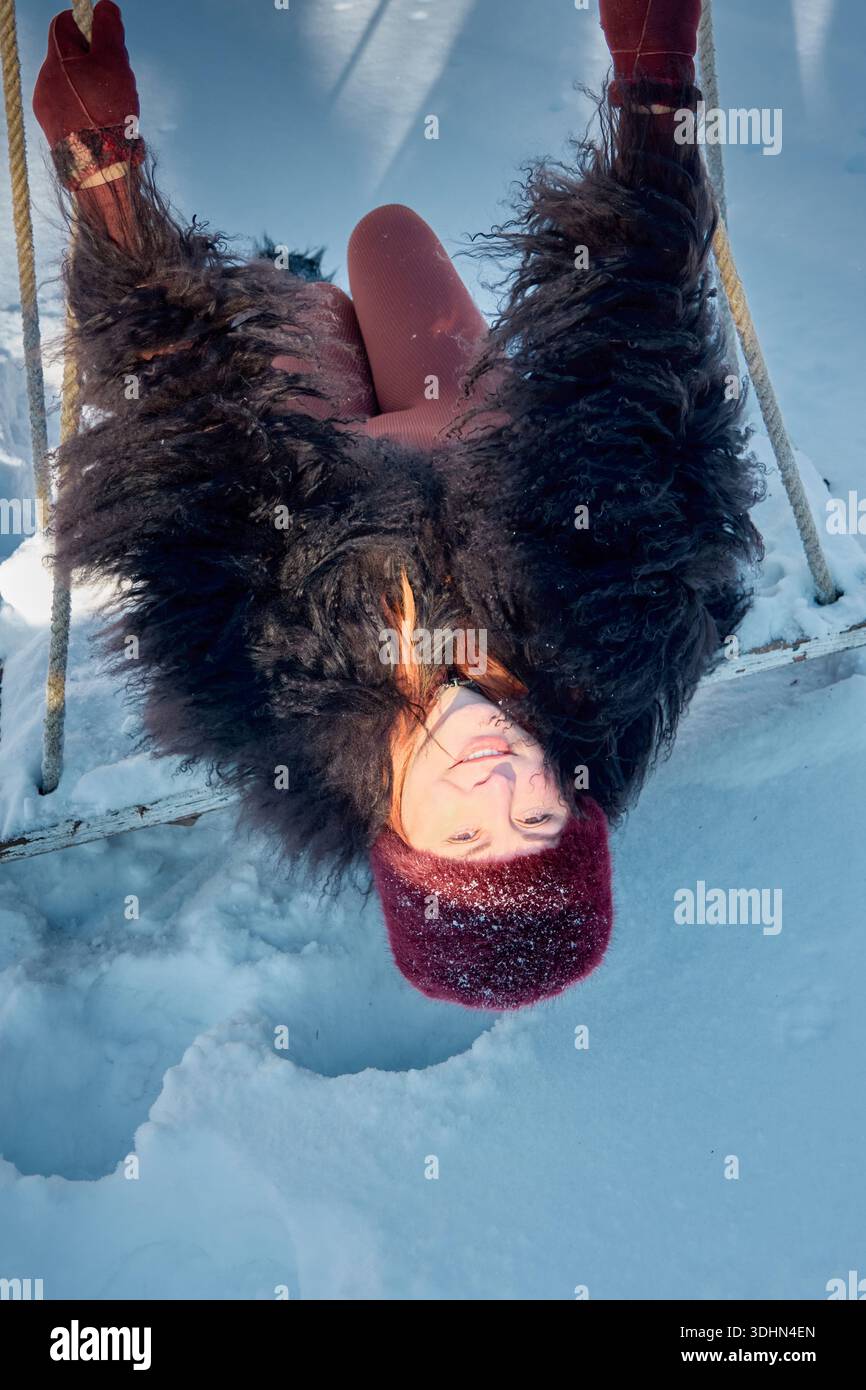 This image captures a cheerful red-haired woman, clad in cozy winter attire, playfully suspended upside down on a swing. The backdrop showcases a sere Stock Photo