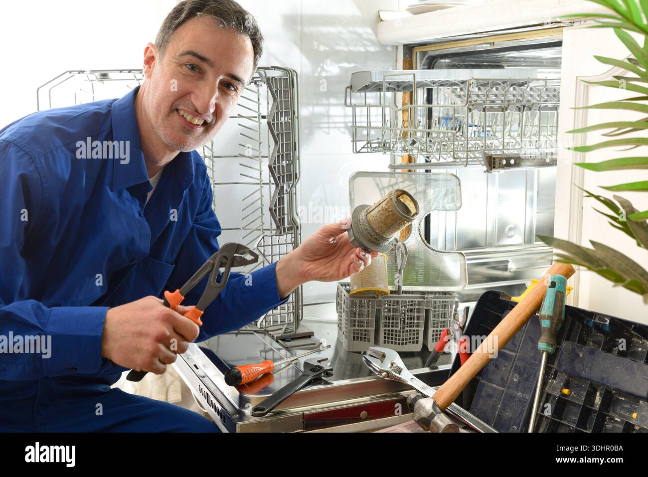 Detail of male technician specialist dressed as a blue working man doing maintenance on a dishwasher with tools around in a white kitchen Stock Photo