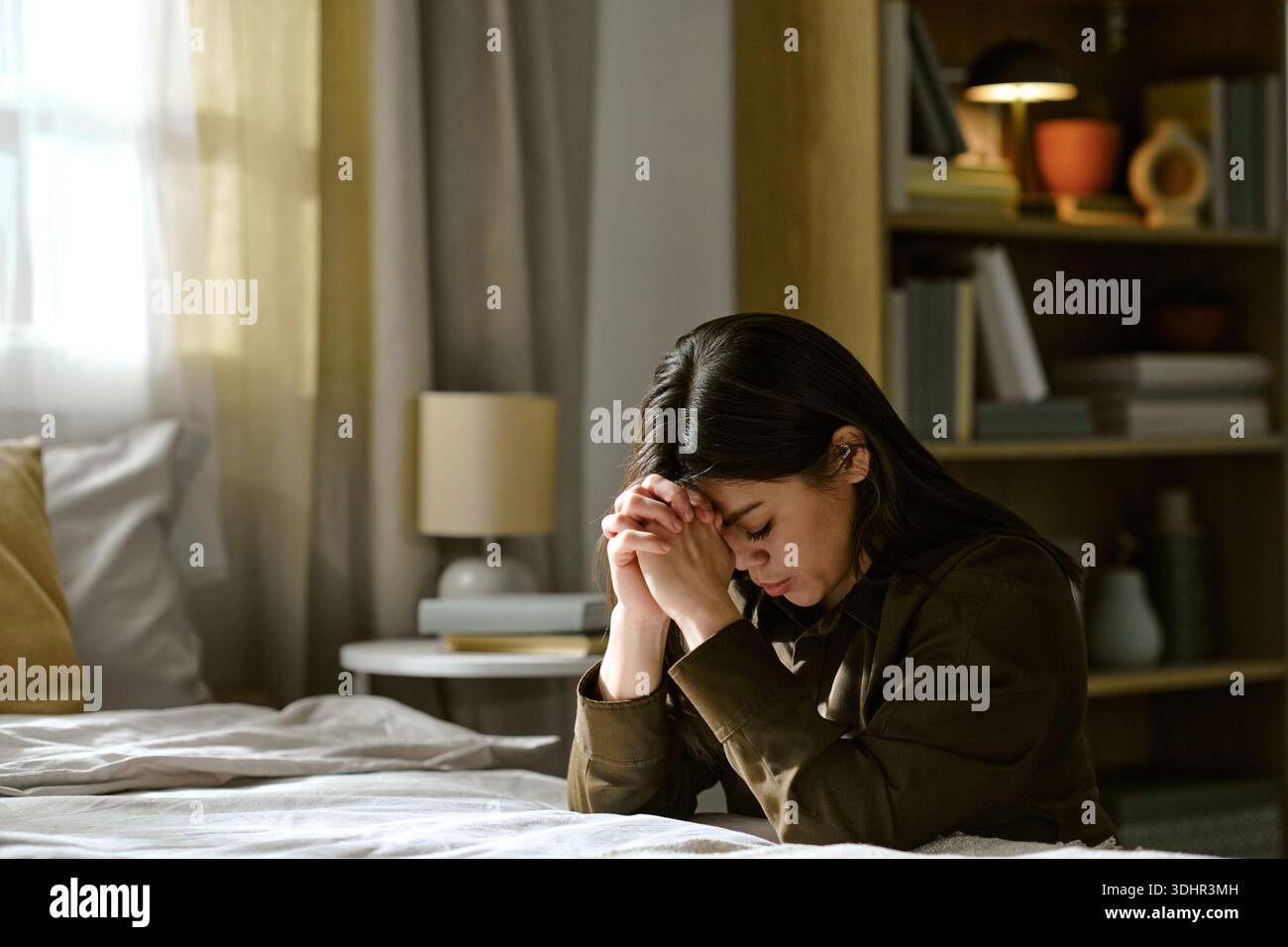 Young Adult Asian Woman Kneeling Beside Bed Bowing Head in Silent Prayer Stock Photo