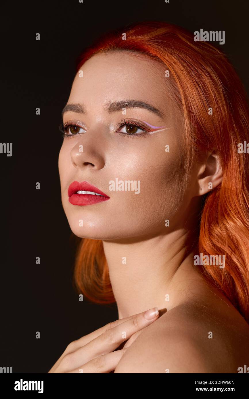 Bright red haired woman showcases contemporary makeup against a dark, artistic background. Stock Photo