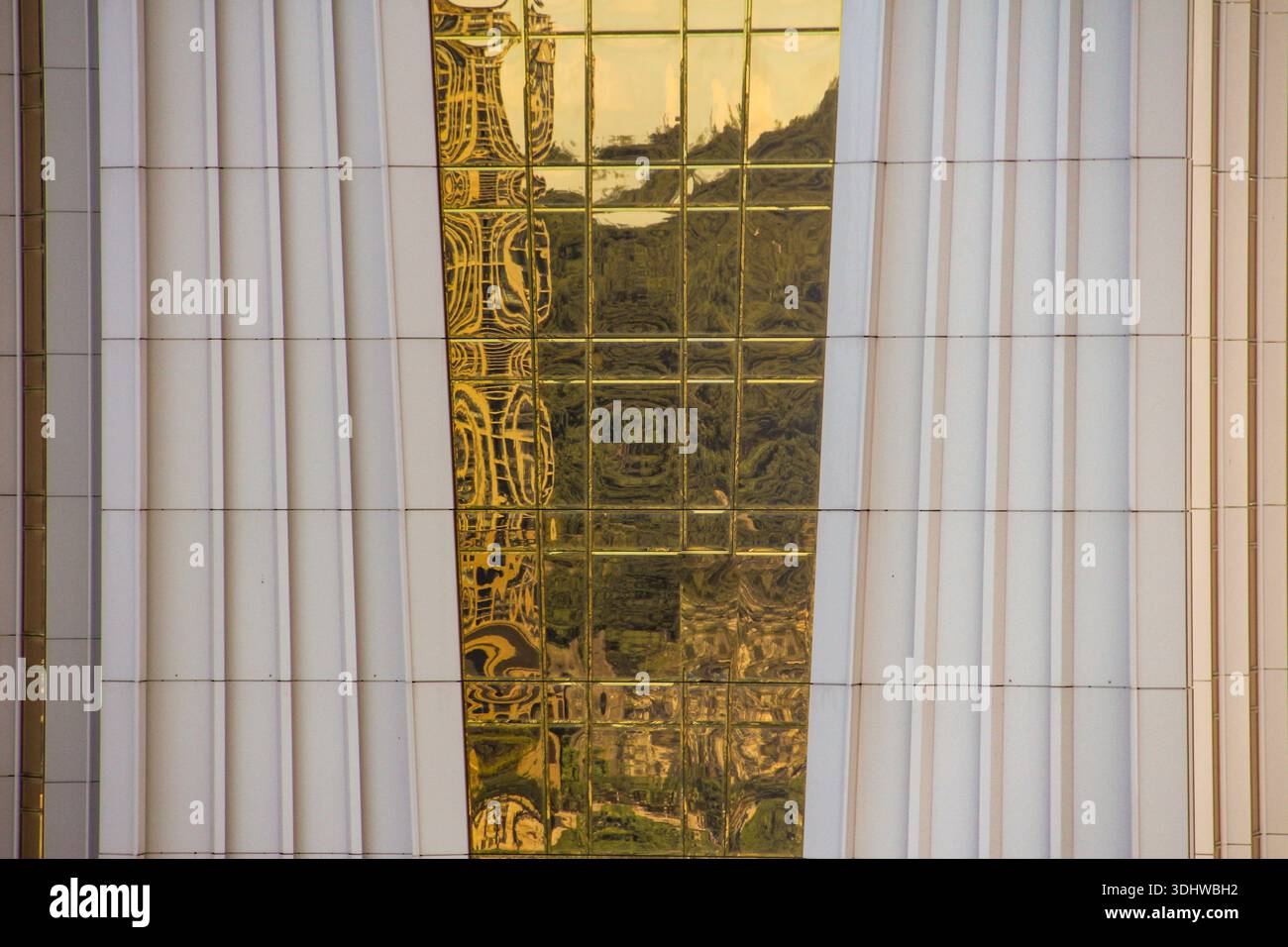 Architectural detail of a modern building featuring a central strip of reflective gold glass panels set between white vertical ribbed pillars. Stock Photo