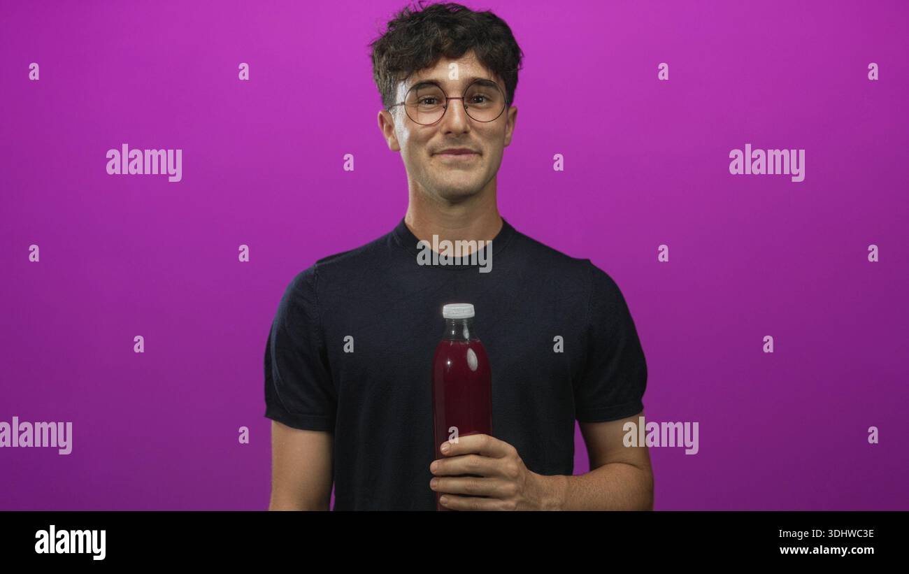 Man young hispanic holding a bottle with hand and cheeks puffed in purple studio wearing glasses and black shirt; playful curiosity. Stock Photo