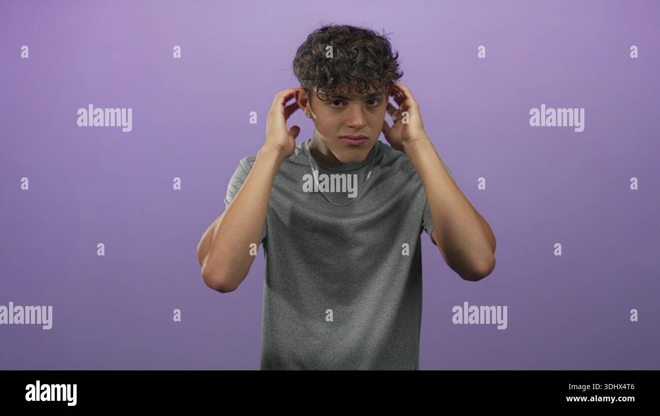 Teenage man touching ears and ruffling hair in purple studio while ...