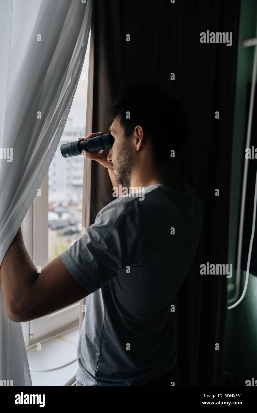 Man curiously peering out window using monocular, watching with intense emotion. Engaging in voyeurism. Observing surveillance. Peeping through Stock Photo