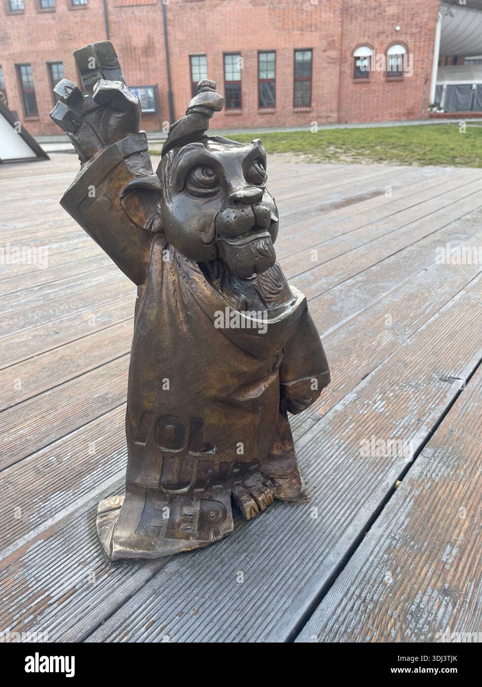 Small bronze bear sculpture standing on a wooden boardwalk, raising its paw in a playful gesture in an urban outdoor setting. - Smartphone Captured Stock Image
