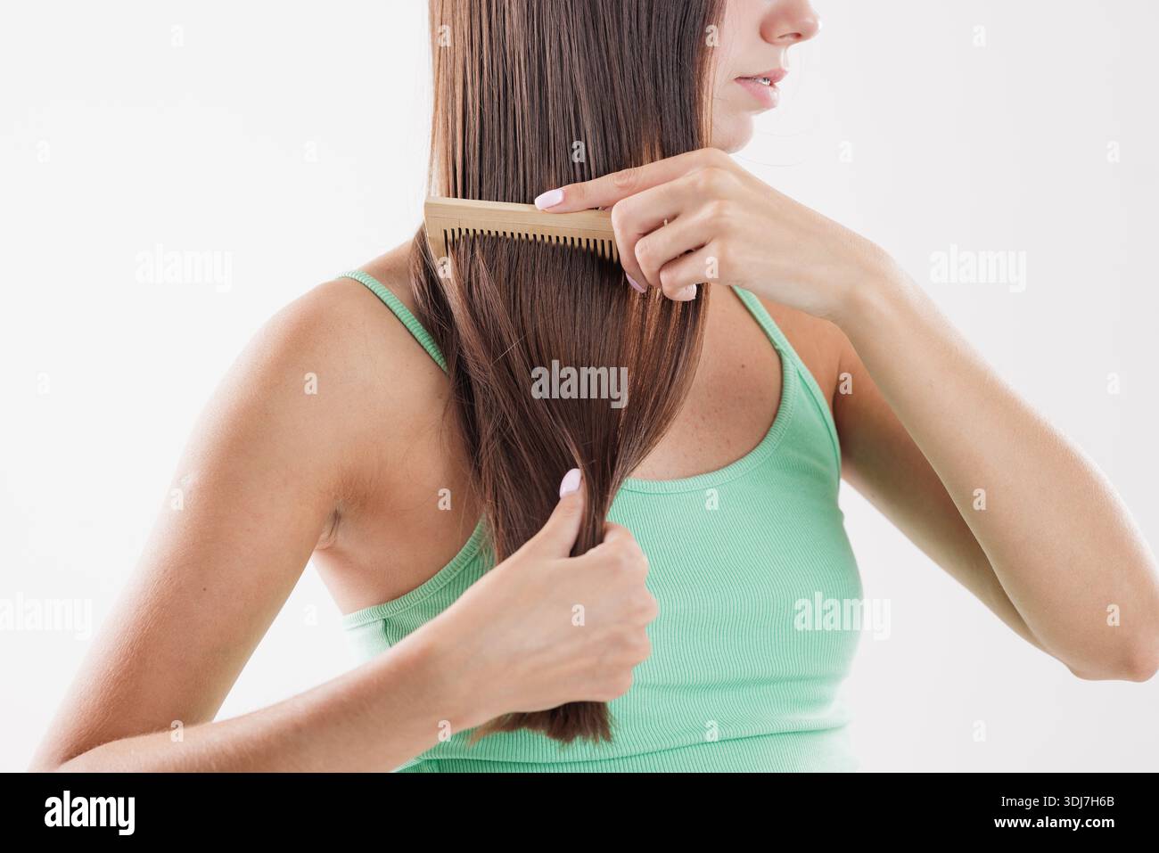 Young woman combing long straight brown hair with a wooden comb, promoting natural beauty, self-care, and healthy hair care routine on a white backgro Stock Photo