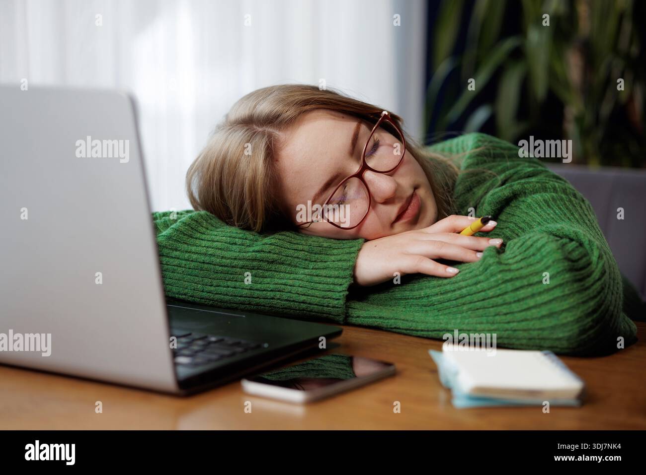 Tired young woman in glasses falling asleep at desk with laptop and notepad, exhausted while working or studying. Stock Photo