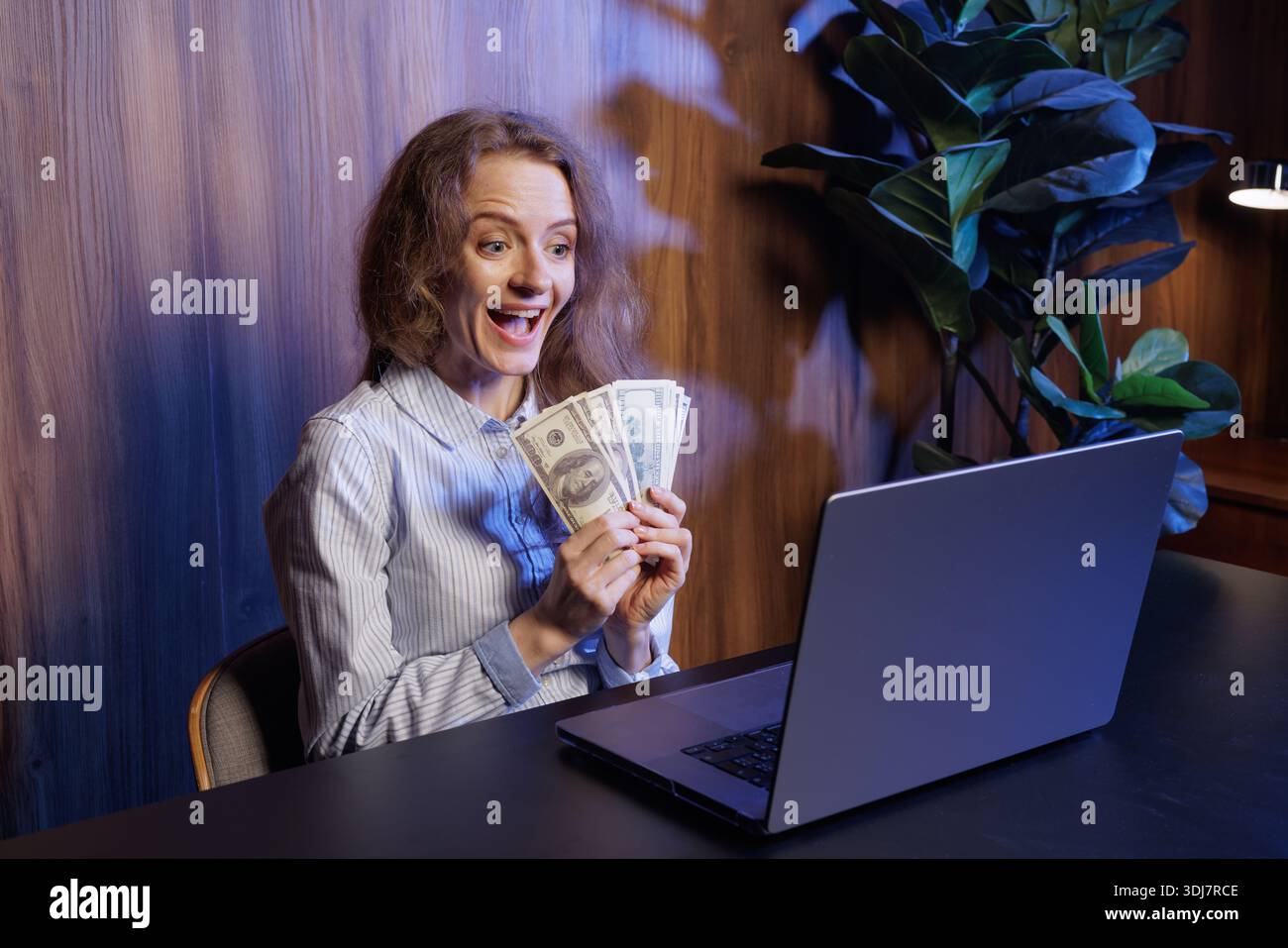 Excited woman holding cash, celebrating online success, winning money, financial achievement, remote work earnings, joyful reaction at laptop Stock Photo