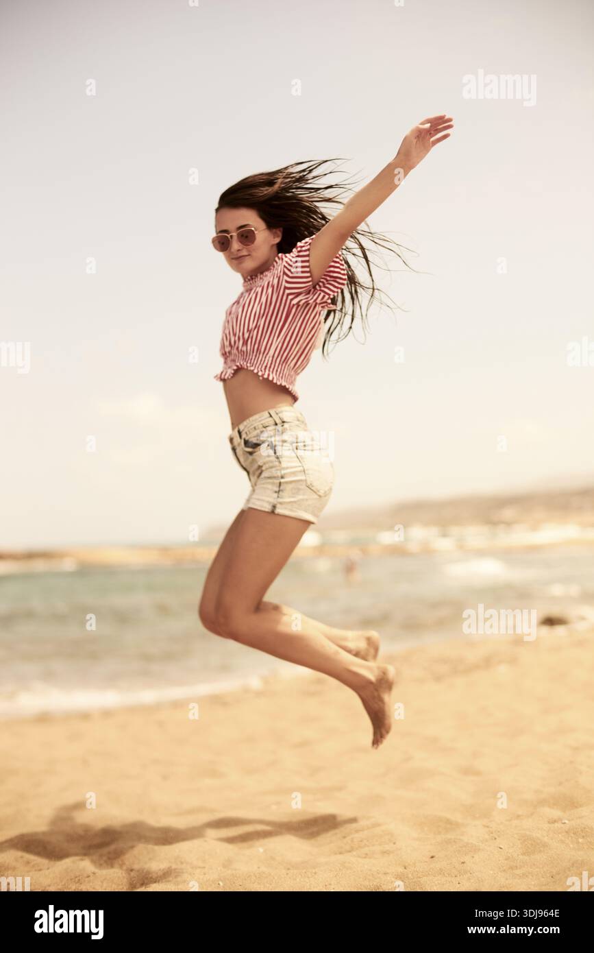 Woman energetically jumping on the beach in sunglasses and casual summer wear, sense of freedom, feeling free, carefree, untroubled, lighthearted, bar Stock Photo