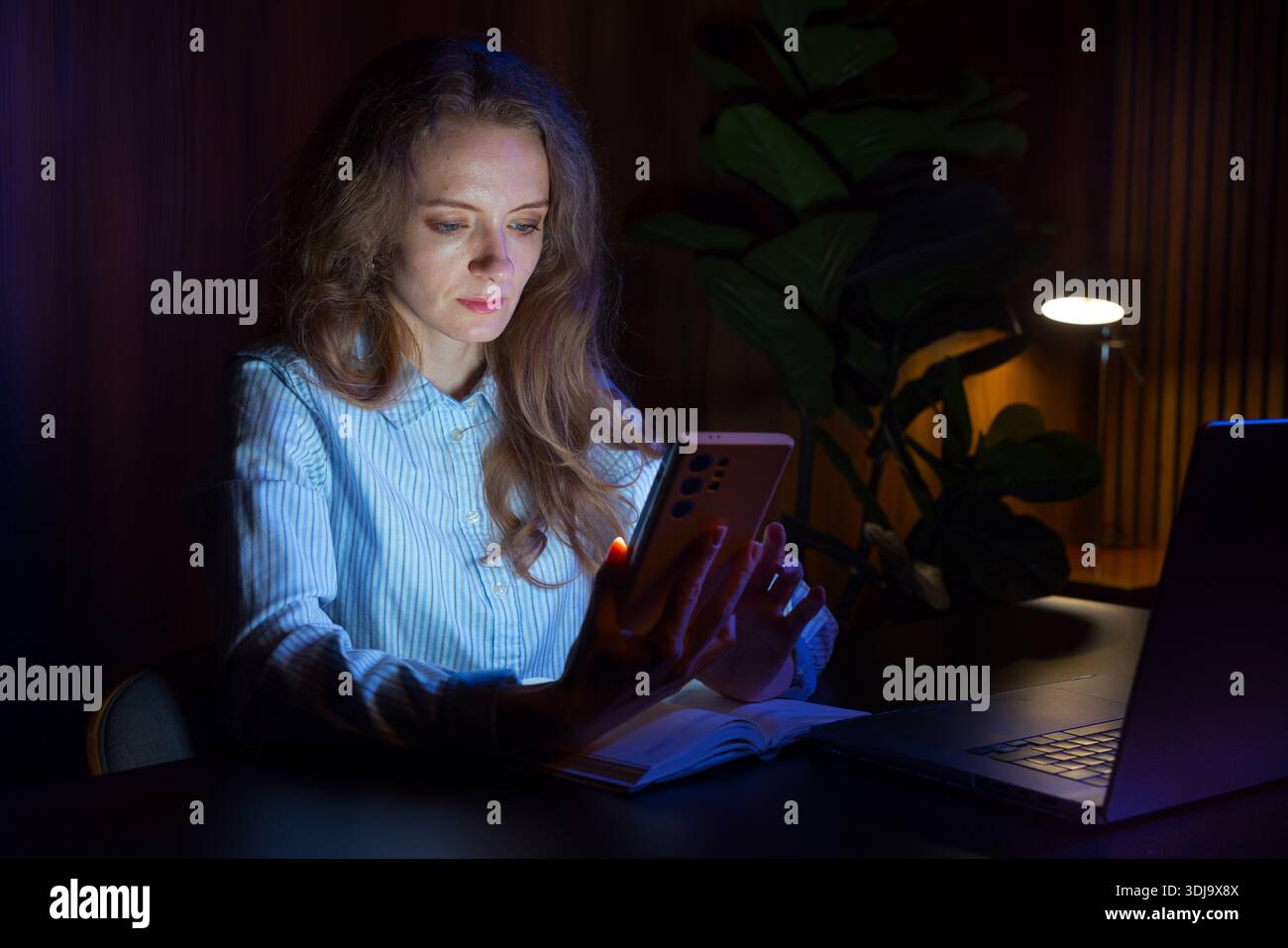 Woman sitting at night on her smartphone, at her desk at home, night work. Stock Photo