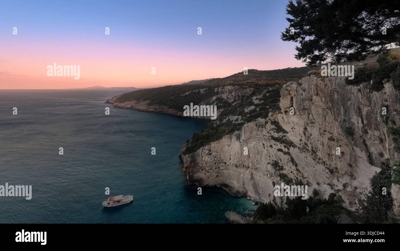 Dramatic aerial view of the Ionian Sea coastline at sunset, featuring towering limestone cliffs adorned with lush green vegetation. A small boat is vi Stock Photo