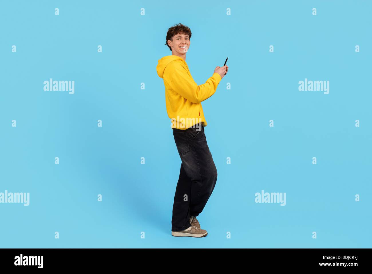 Smiling young man in yellow hoodie holding smartphone with surprised expression Stock Photo