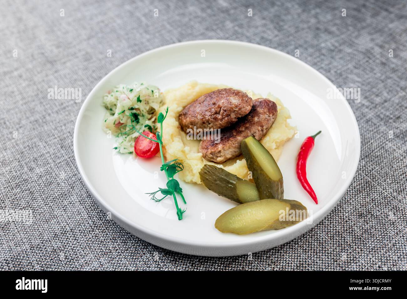 Wild boar cutlets with mashed potatoes. Served on a white plate with pickled cucumbers and sauerkraut. Decorated with herbs, tomatoes, red chili peppe Stock Photo