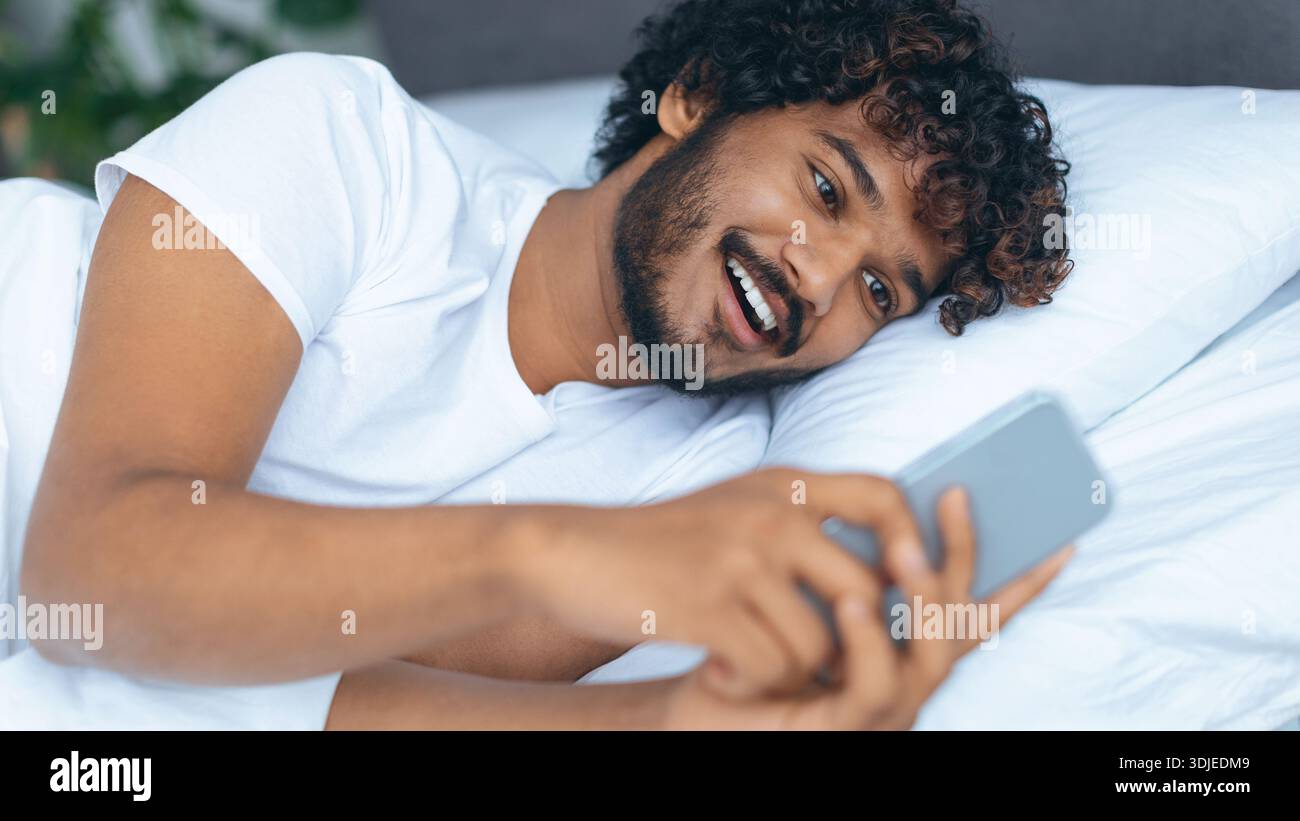 Man Lying on Bed Looking at Smartphone and Smiling While Enjoying Content Stock Photo