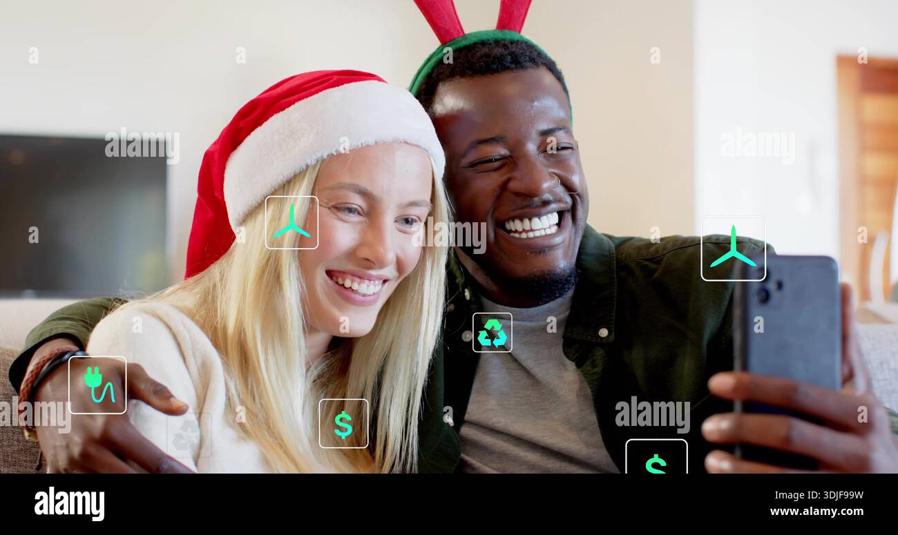 Smiling adult couple taking selfie in living room, with smartphone, Santa hat, antlers, green icons Stock Photo
