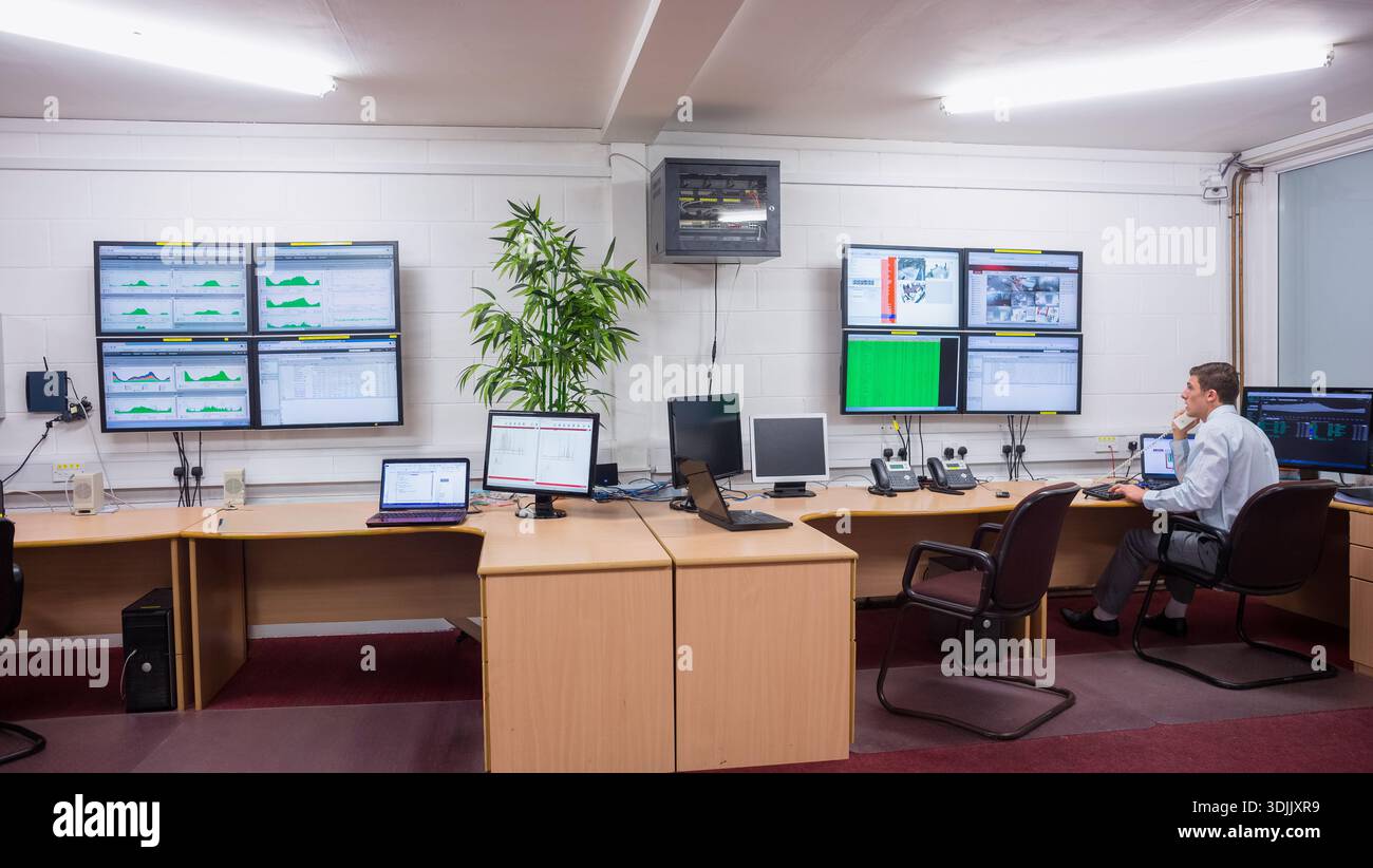 Male operator wearing shirt sitting at desk in control room monitoring screens cables, copy space. Dashboards, displays, monitors, laptops, plant, car Stock Photo