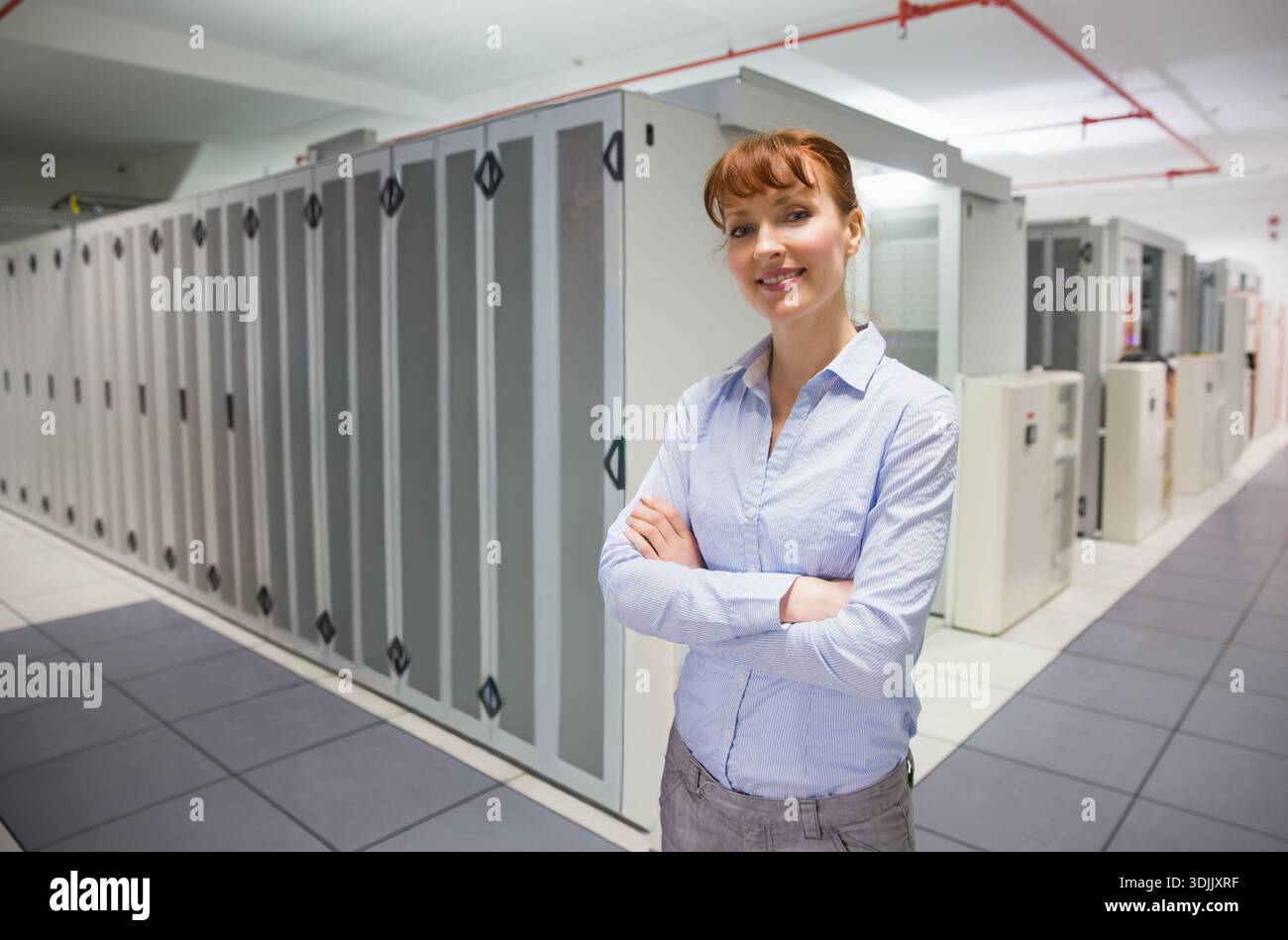Female technician standing arms crossed in blue shirt gray pants at data center racks, copy space. Serverroom, cabinet, corridor, cooling, electrical, Stock Photo