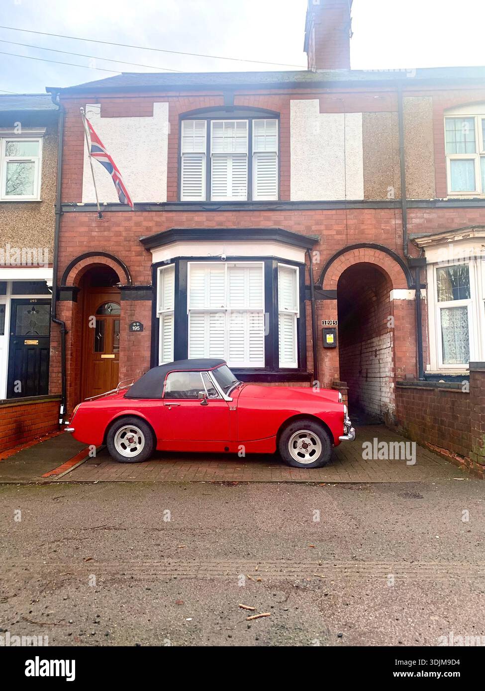 A classic red convertible parked outside a traditional British brick townhouse complete with bay window and union jack flag creating a street scene - Smartphone Captured Stock Image