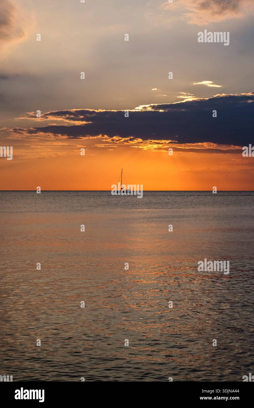 Sunset over the sea in Porec. A lone sailboat is visible on the calm water against a glowing orange sky with dark clouds. Stock Photo