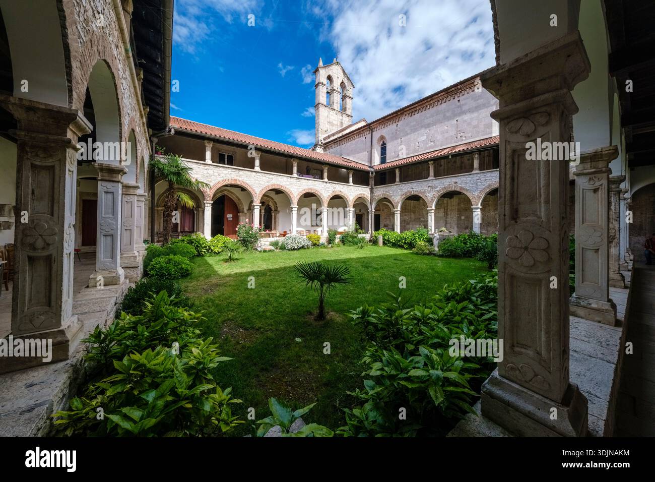 Historic Franciscan Monastery cloister in Pula with a green courtyard, stone arches, and a bell gable rising into a blue sky. Stock Photo