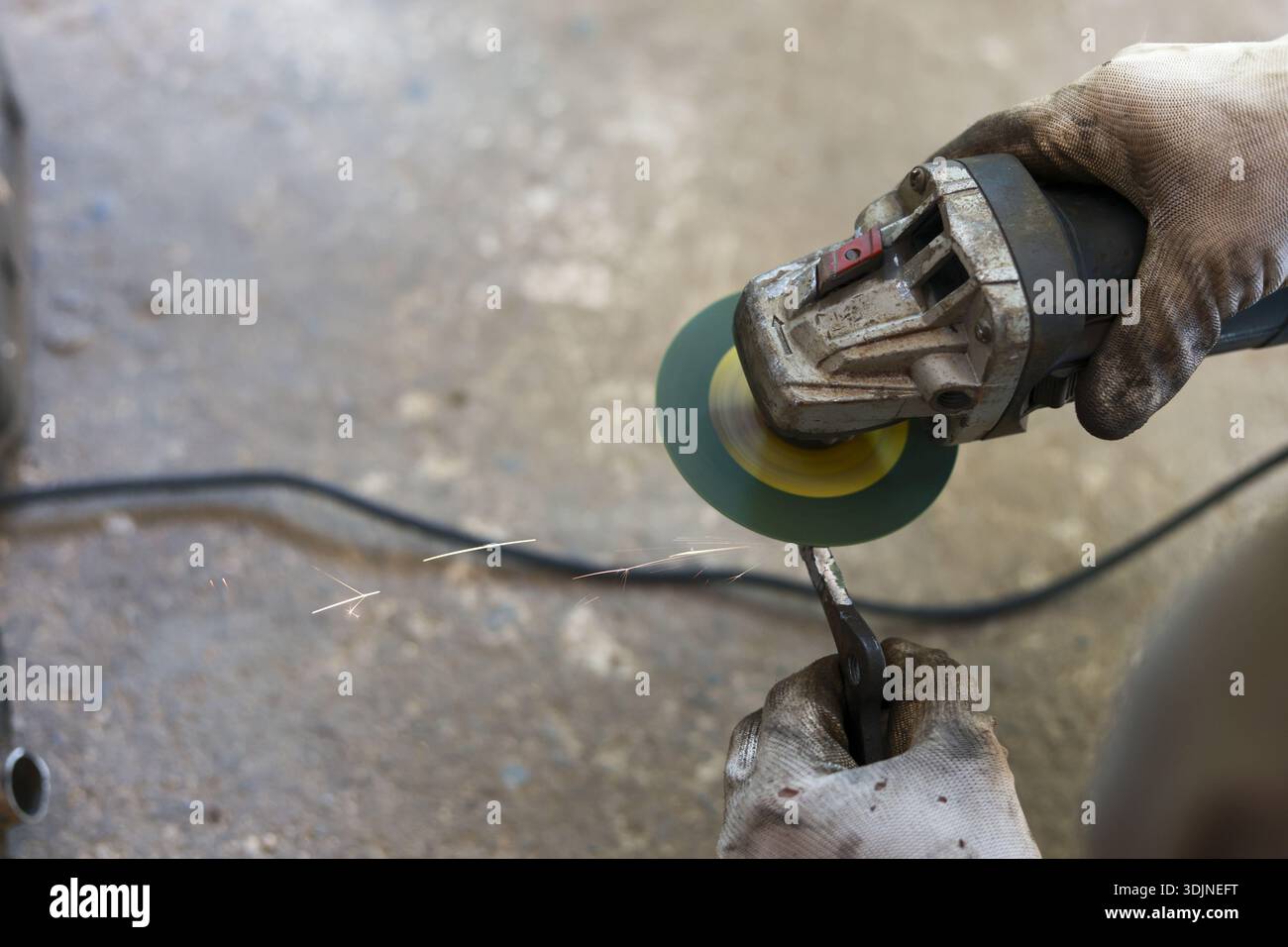 Focused worker in workshop grinds piece of metal with an angle grinder. Dirty gloved hands hold powerful tool and object, process creating sparks on c Stock Photo