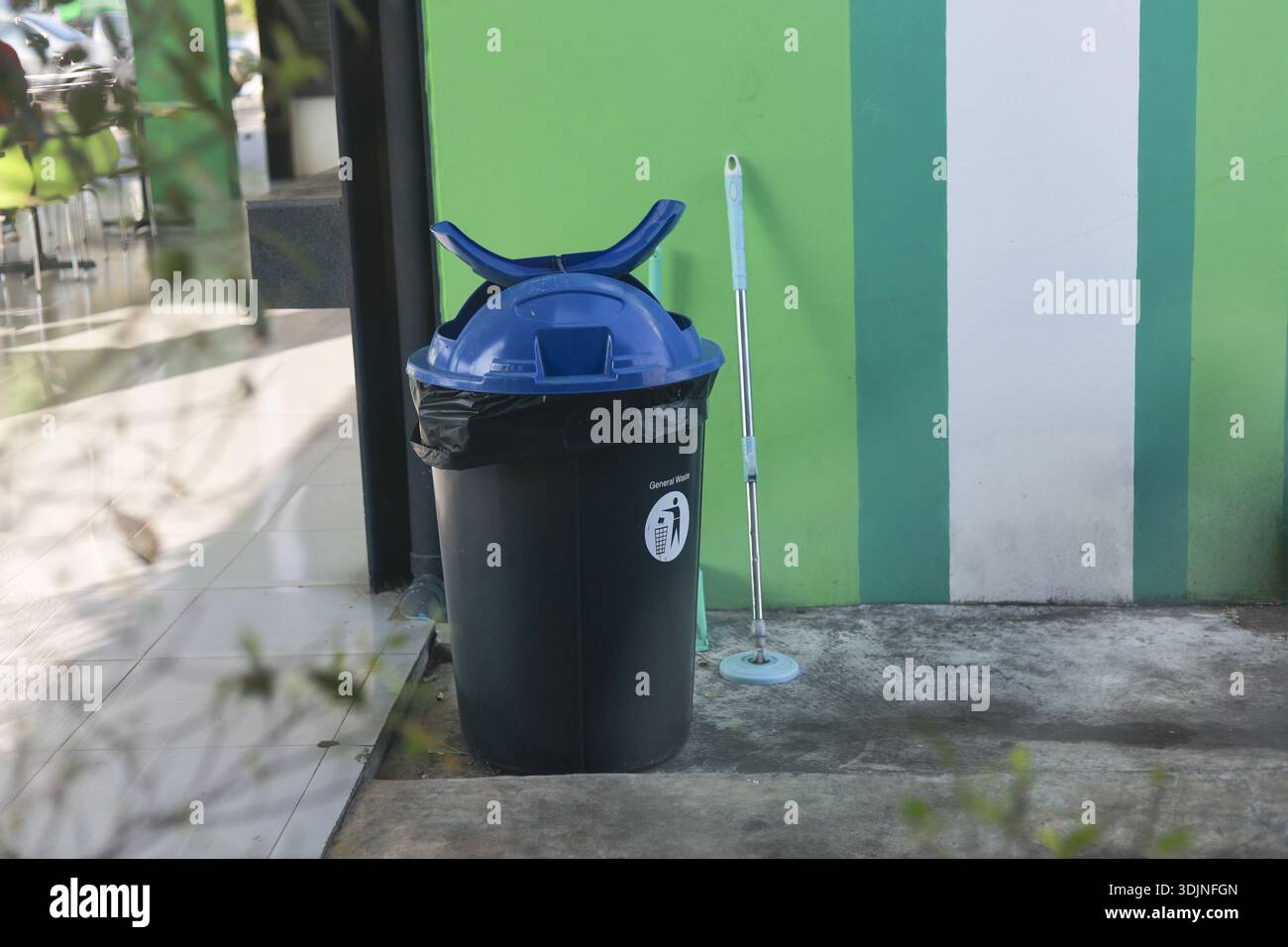 Black trash can with blue lid on concrete floor. This outdoor setting shows mundane scene with green striped wall and leaning mop for cleaning public Stock Photo