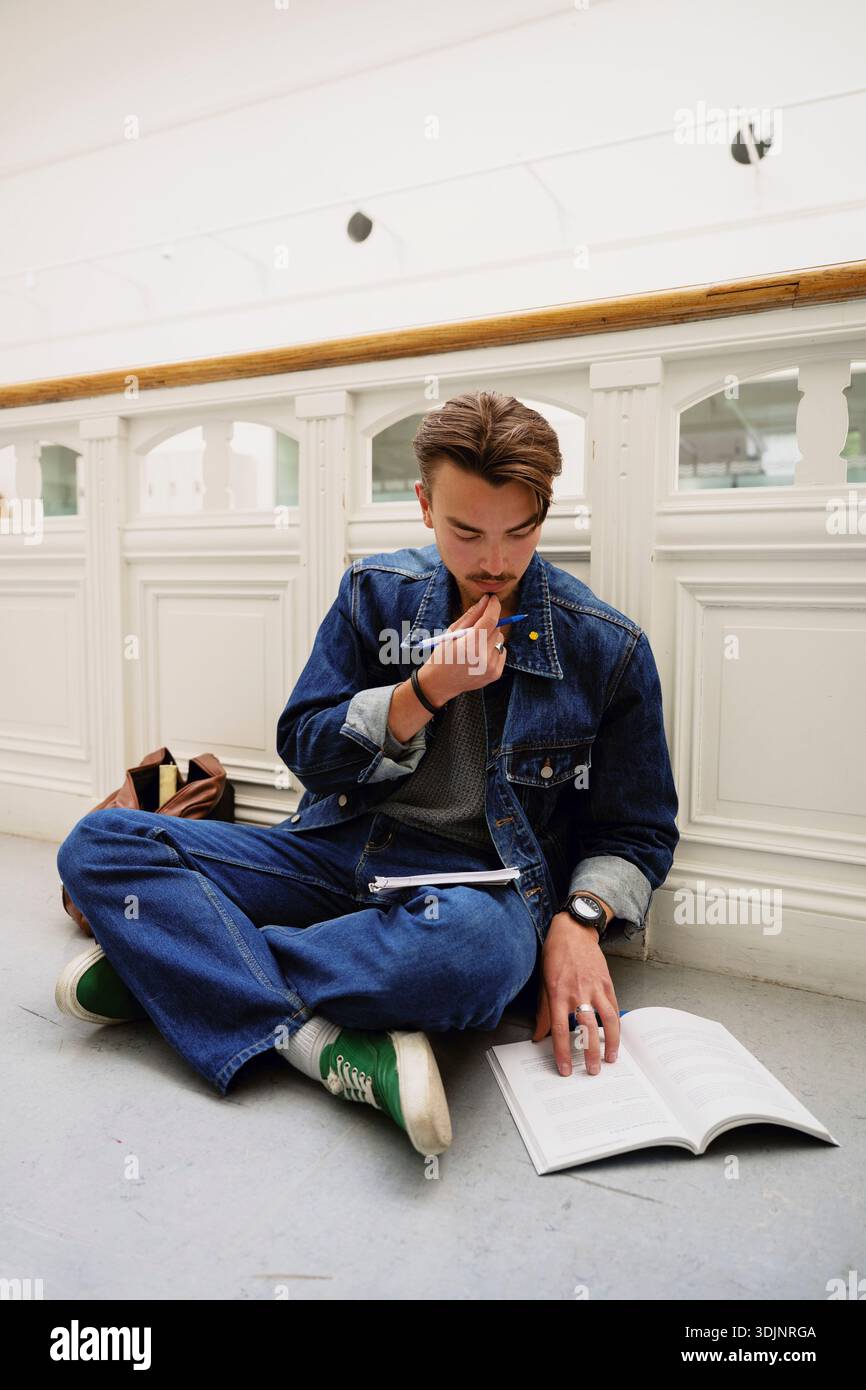 Focused young male student studying while sitting cross-legged on ground in corridor at college Stock Photo